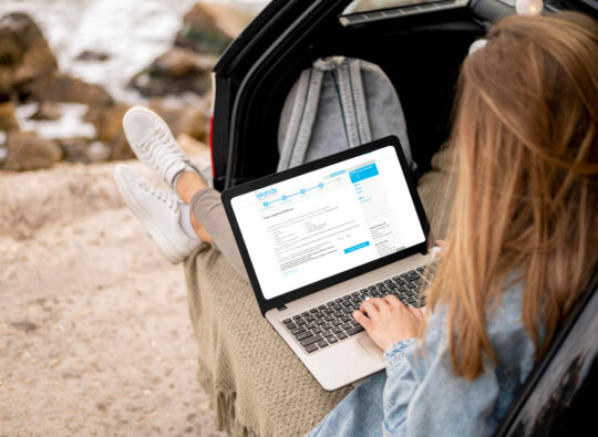 Woman relaxing in her car boot while using a laptop displaying the Islands Insurance travel insurance medical screening webpage, planning for her journey with peace of mind.